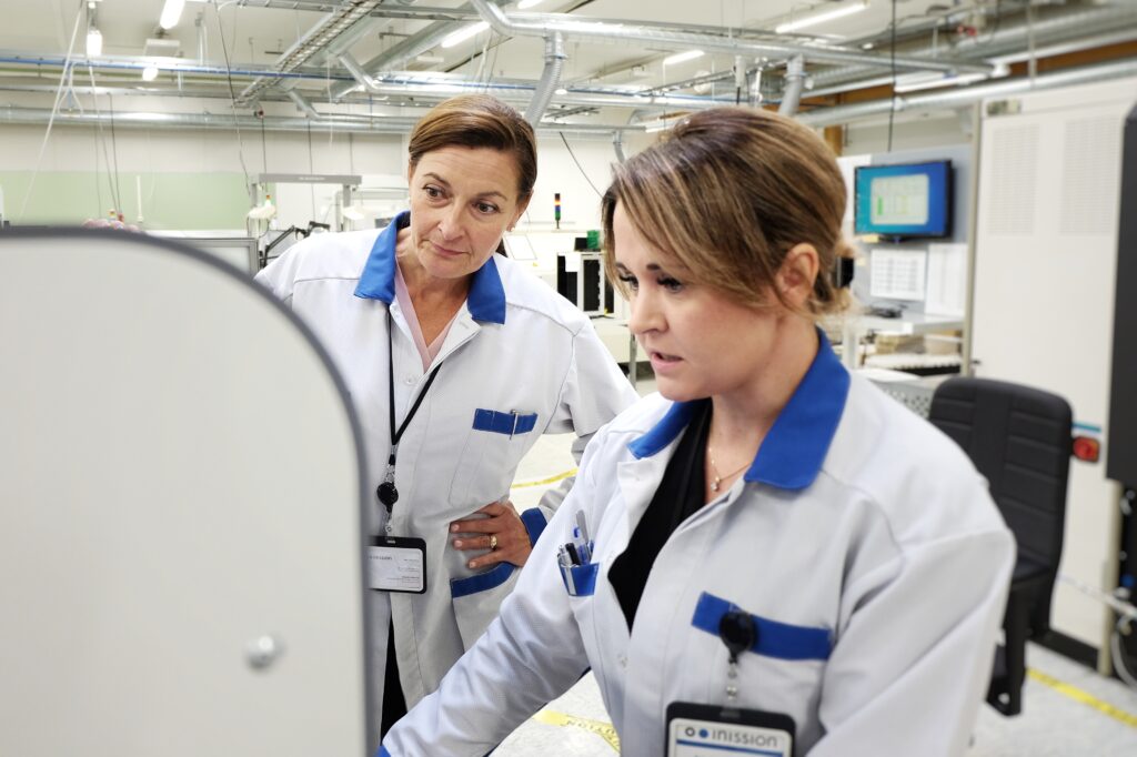 Two female employees wearing white and blue lab coats are working together in an electronics contract manufacturing facility, focused on operating a machine. The background shows a clean industrial environment with electronics production equipment, wiring, and computer monitors.